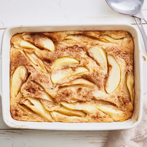 A warm slice of baked apple cake sits on a white plate, topped with crème fraîche and a dusting of powdered sugar.